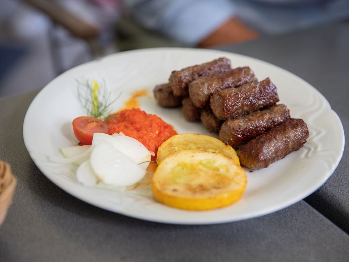 Čevapčići na tanjuru s ajvarom, sirovim lukom, rajčicom i pečenom tikvicom.