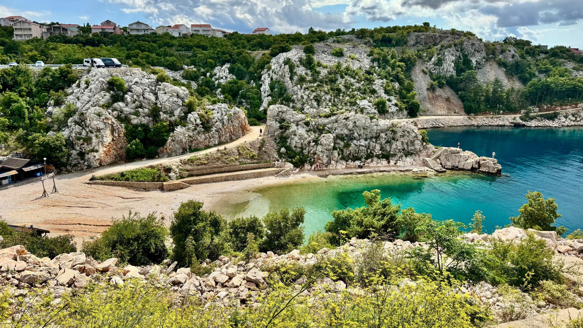 Uvala u Karlobagu: bijele krške stijene, tirkizno more i obalna cesta iznad plaže.